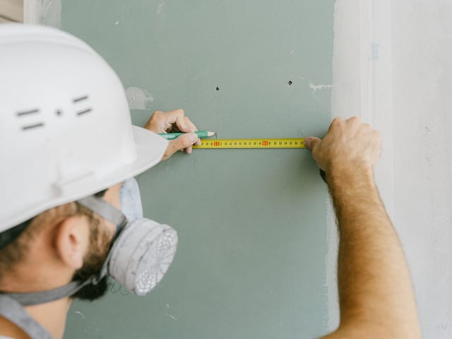Handwerker mit Helm misst eine Gipskartonplatte mit dem Bandmaß aus