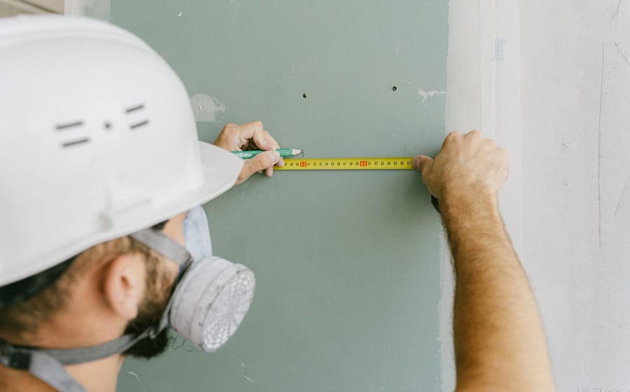 Handwerker mit Helm misst eine Gipskartonplatte mit dem Bandmaß aus